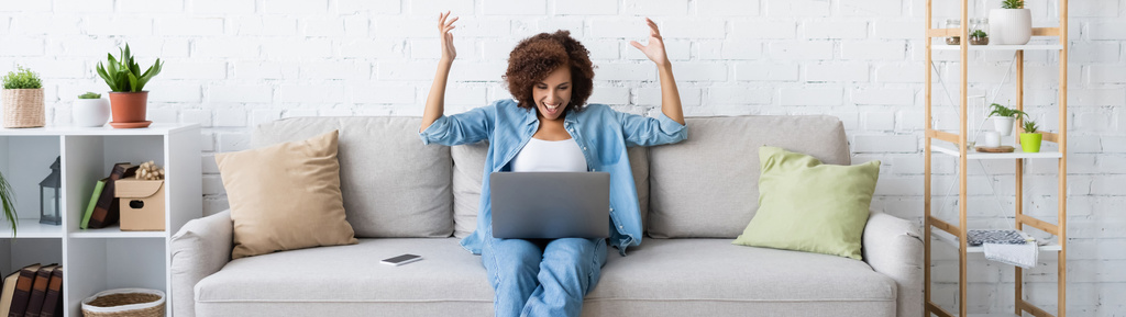 animado afro-americano mulher usando laptop enquanto sentado no sofá e trabalhando em casa, banner - Foto, Imagem