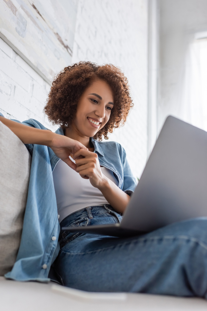 vue à faible angle de femme afro-américaine heureuse en utilisant un ordinateur portable tout en étant assis sur le canapé  - Photo, image