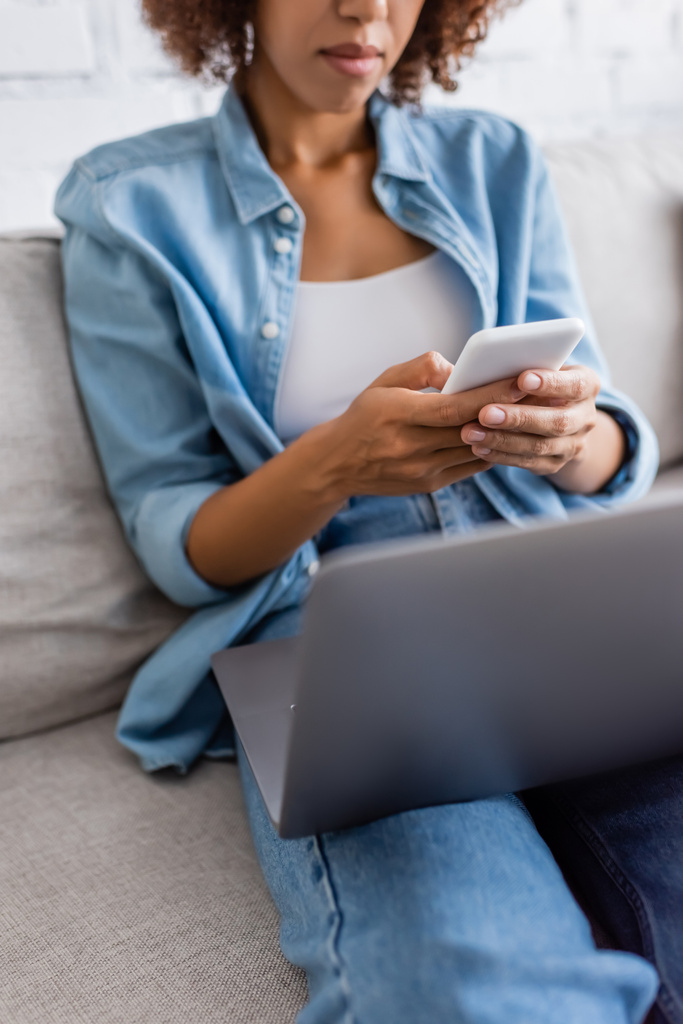 przycięty widok afrykańskiej amerykańskiej kobiety wiadomości na smartfonie w pobliżu laptopa podczas siedzenia na kanapie  - Zdjęcie, obraz