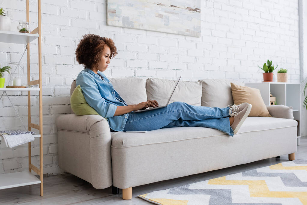 ソファに座って自宅で仕事をしながらノートパソコンを使うアフリカ系アメリカ人女性に焦点を当て  - 写真・画像