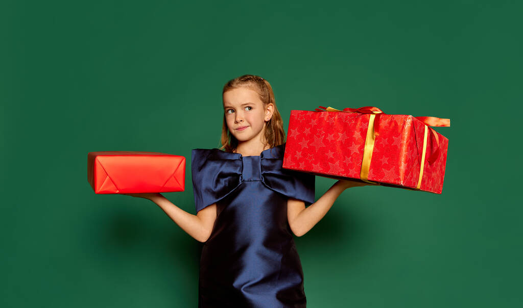 Portrait of little girl, child in dress posing with presents box isolated over green studio background. Thoughtful. Concept of childhood, emotions, facial expression, lifestyle, New Year, Christmas - Photo, Image
