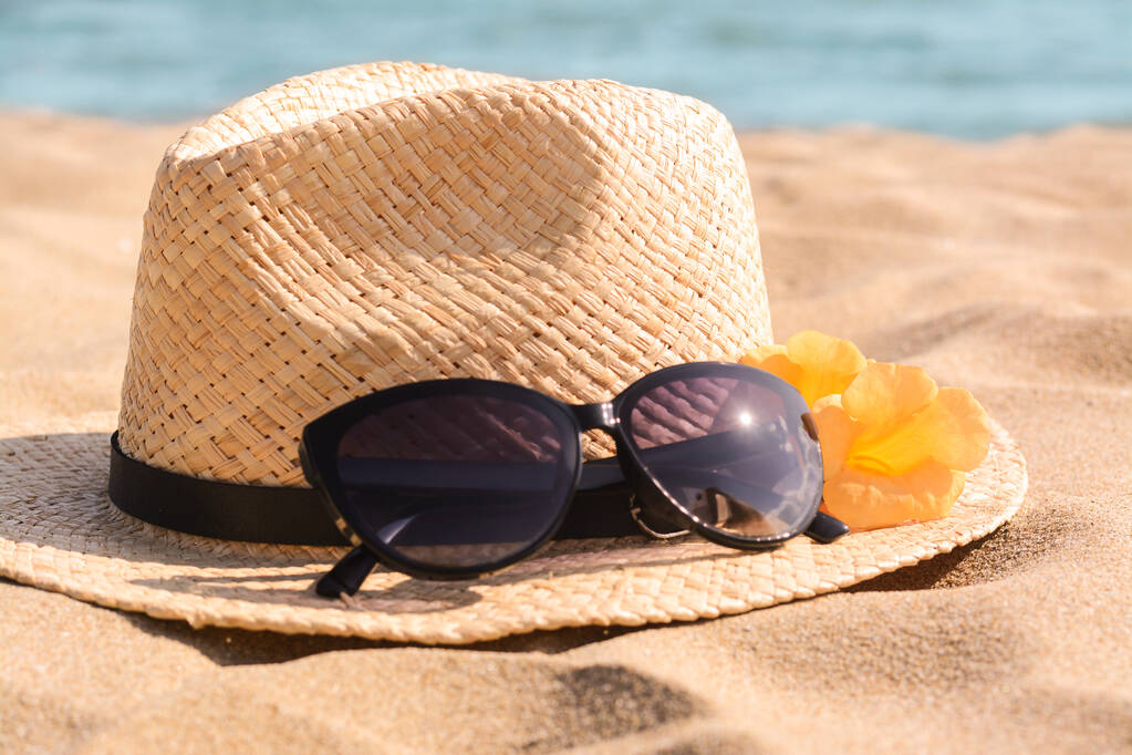 Καπέλο με όμορφα γυαλιά ηλίου και λουλούδια στην άμμο κοντά στη θάλασσα, closeup - Φωτογραφία, εικόνα