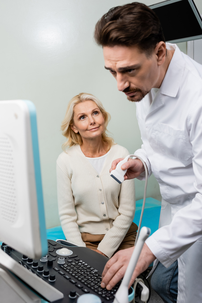 attentive doctor looking at monitor of ultrasound machine near smiling middle aged woman - Photo, Image