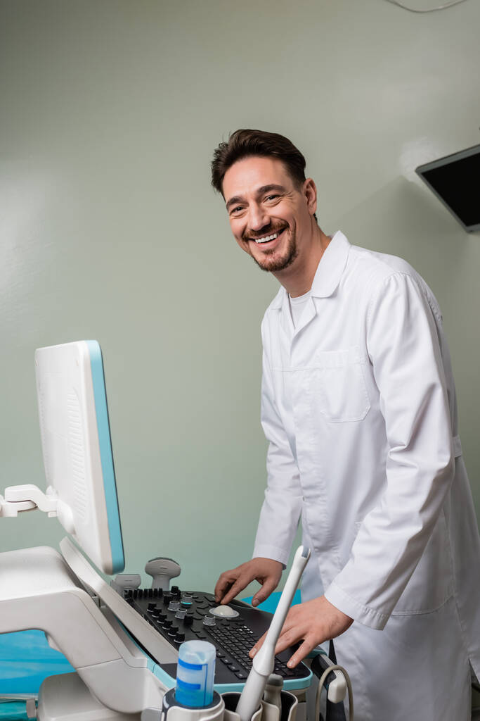 médico feliz en bata blanca mirando a la cámara mientras opera la máquina de ultrasonido en la clínica - Foto, imagen