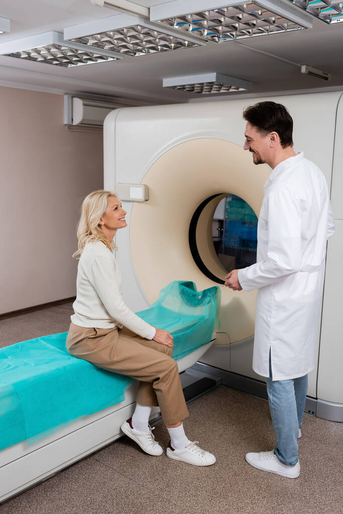 radiologo in camice bianco che parla con una donna matura sorridente seduta vicino alla macchina tomografica computerizzata - Foto, immagini