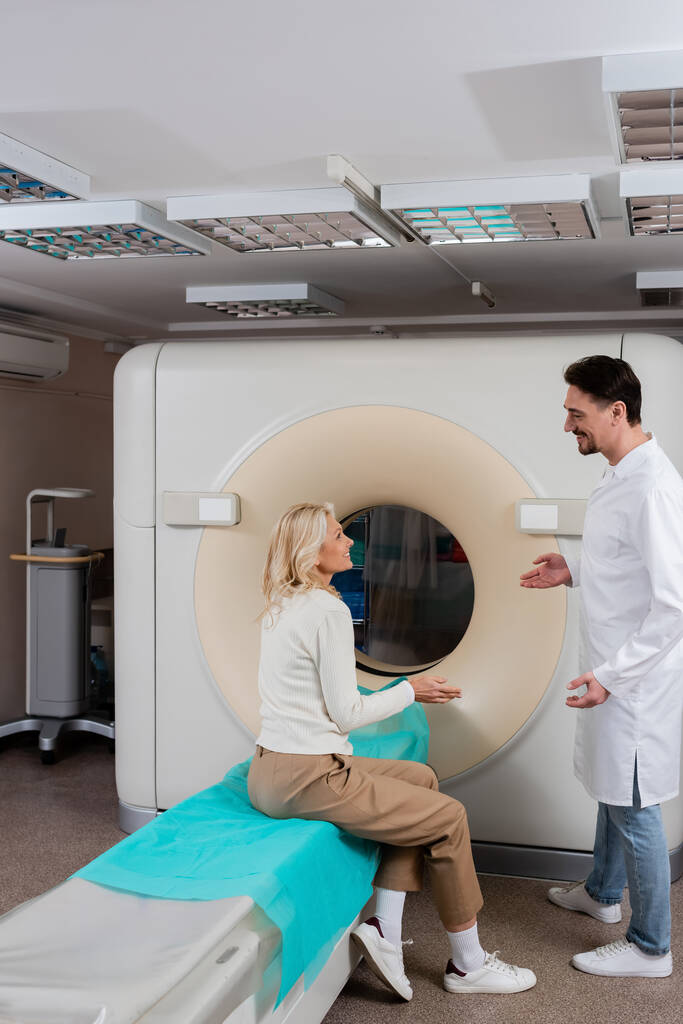 вид збоку позитивного лікаря і пацієнта розмовляє і жестикулює біля комп'ютерного томографічного сканера в клініці
 - Фото, зображення