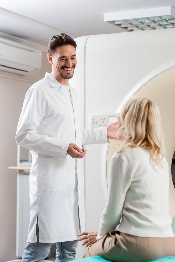 médecin joyeux en manteau blanc parlant à la femme blonde près de la machine de tomographie informatique - Photo, image