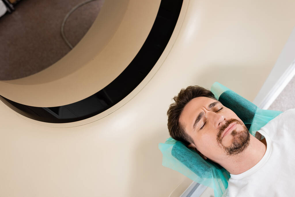 Yüksek açı görüşlü, gözleri kapalı esmer bir adam bilgisayarlı tomografi tarayıcısında tarama yapıyor. - Fotoğraf, Görsel