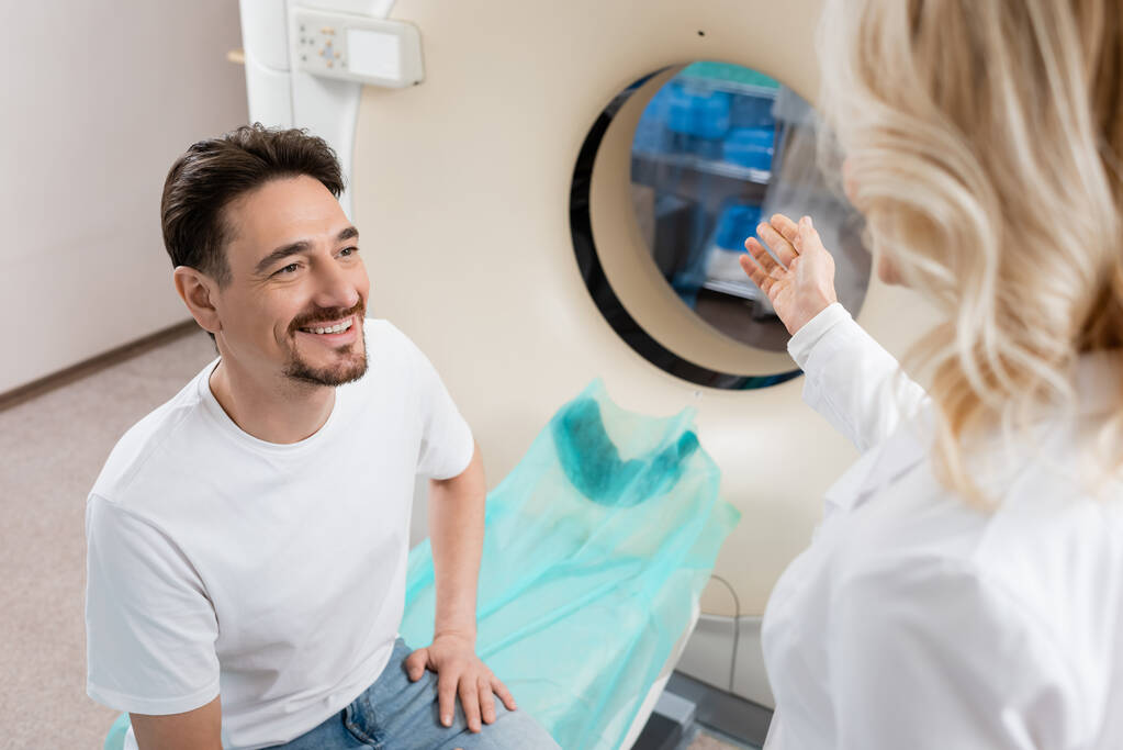 verschwommener Arzt zeigt in der Nähe lächelnder Patientin auf Computertomographiegerät - Foto, Bild