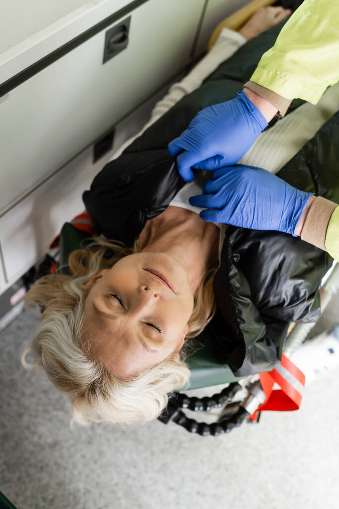 Widok z góry na ratownik medyczny zdejmujący kurtkę od nieprzytomnej kobiety w średnim wieku w pojeździe ratunkowym  - Zdjęcie, obraz