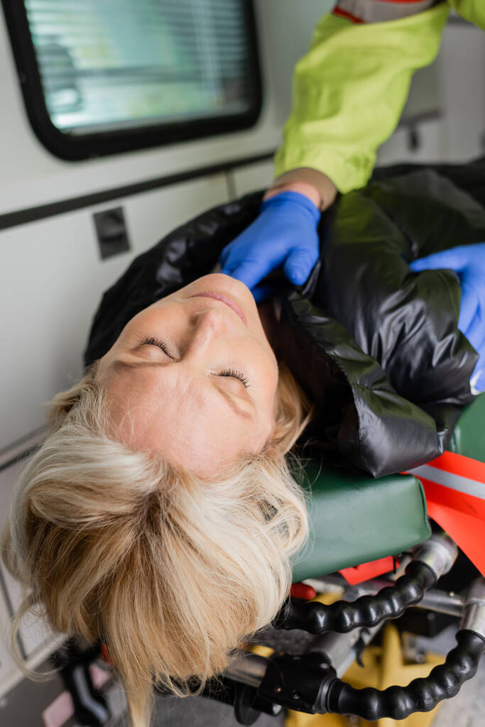 Bewusstlose Frau liegt neben verschwommenem Sanitäter in Latexhandschuhen im Einsatzfahrzeug  - Foto, Bild