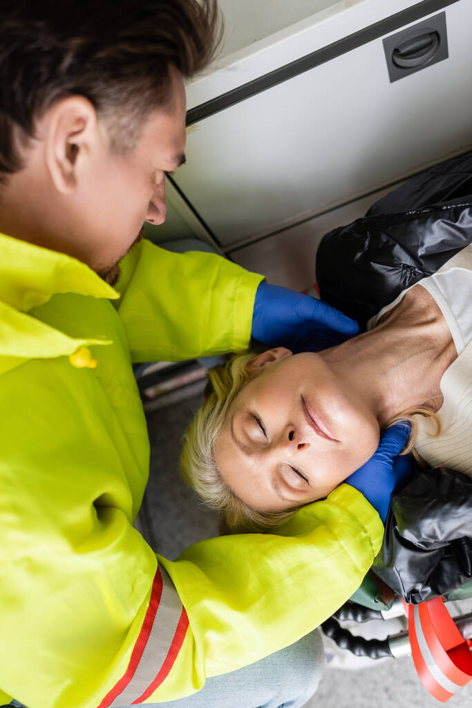 Vista superior de paramédico en guantes de látex comprobando cuello de mujer de mediana edad en vehículo de emergencia  - Foto, Imagen