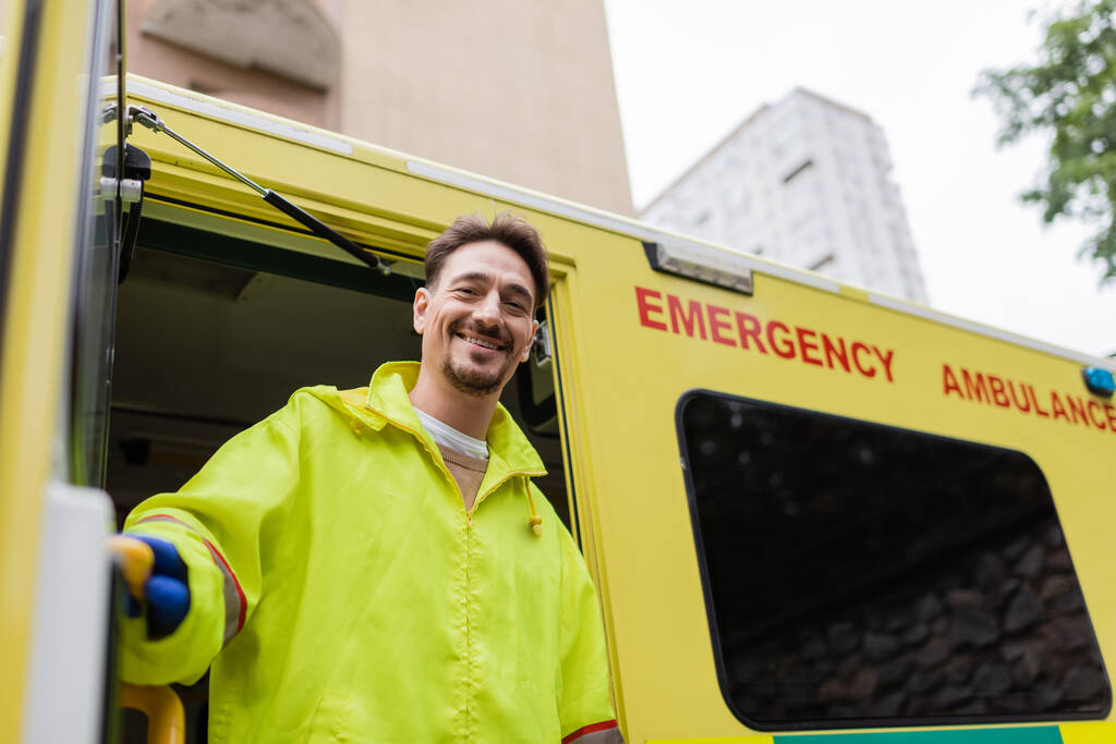 Sorridente paramedico guardando la fotocamera vicino al veicolo ambulanza con porta aperta sulla strada  - Foto, immagini