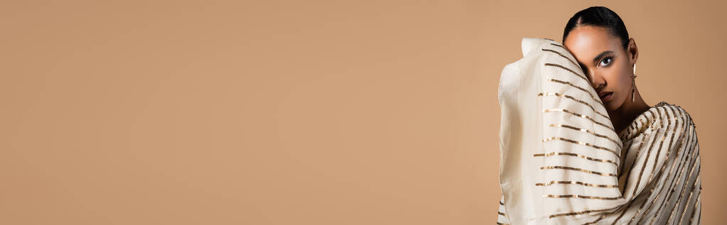 elegantti afrikkalainen amerikkalainen nainen kultainen korvakoru katselee kameraa poseeratessaan eristetty beige, banneri  - Valokuva, kuva