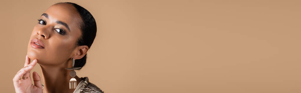 элегантная африканская американка в золотых украшениях касаясь подбородка изолированы на бежевый, баннер  - Фото, изображение