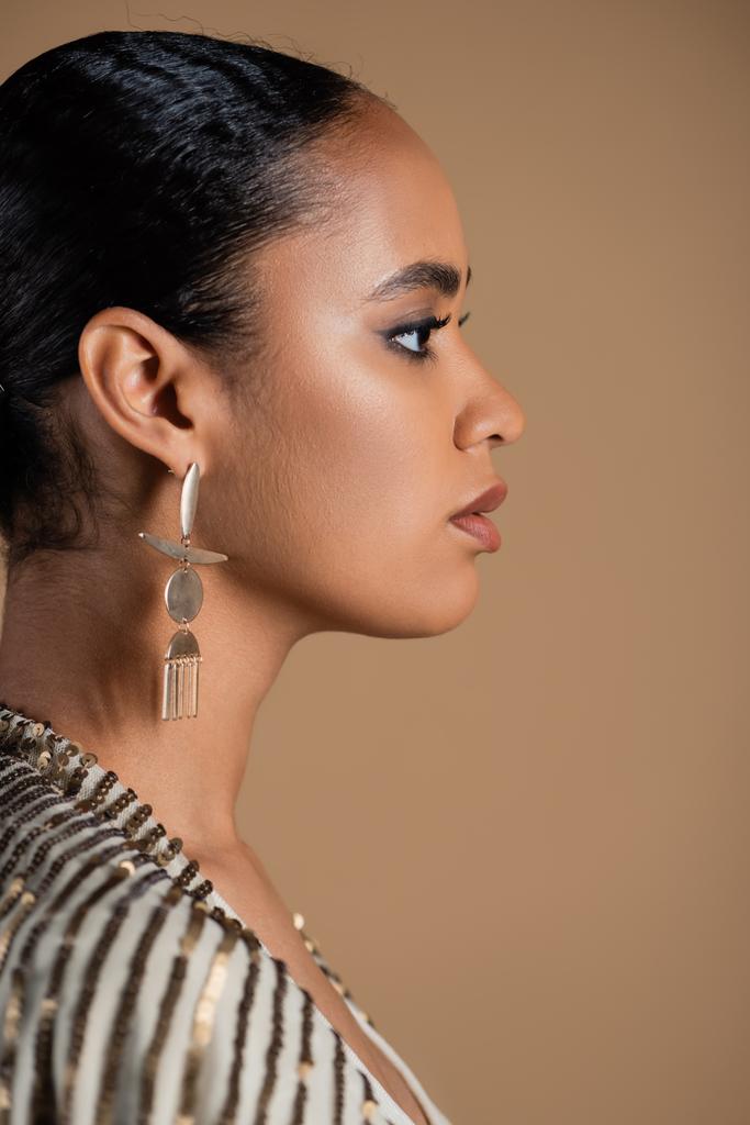 πλαϊνή άποψη της νεαρής Αφροαμερικανής γυναίκας σε πολυτελή χρυσά κοσμήματα που απομονώνονται σε μπεζ  - Φωτογραφία, εικόνα