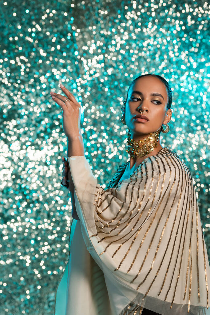 piercing afro-amerikai modell arany fólia a nyakon pózol, és félrenéz fényes kék háttér  - Fotó, kép