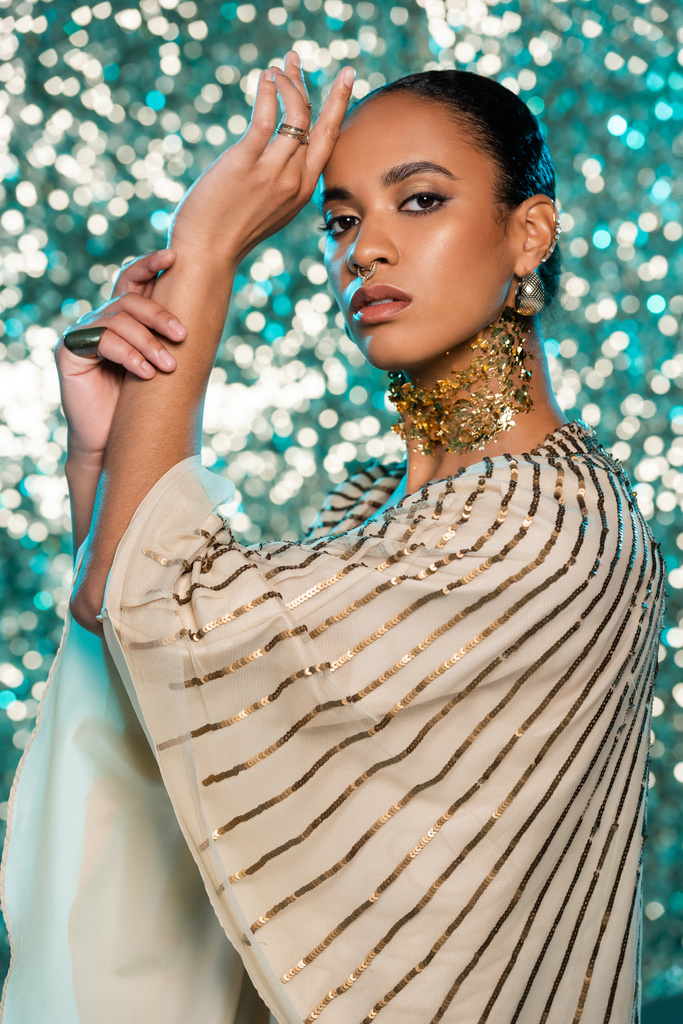 Parlak mavi arka plandaki kameraya bakarken boynunda altın folyo ve mücevher olan delinmiş Afro-Amerikan modeli.  - Fotoğraf, Görsel