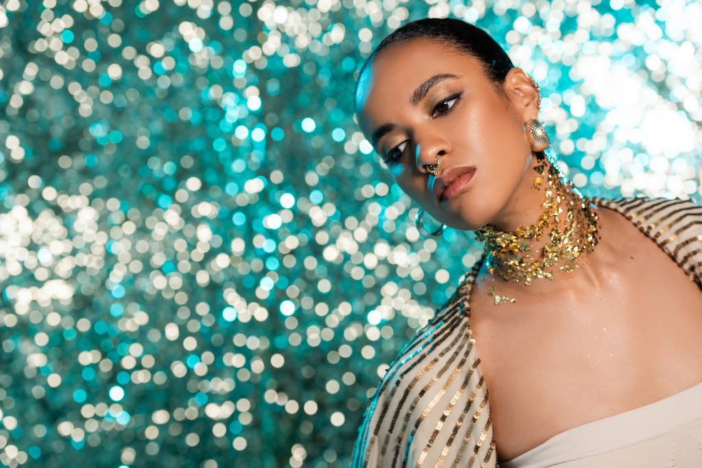 african american model with piercing and gold on neck posing while looking away on shiny blue background  - Photo, Image