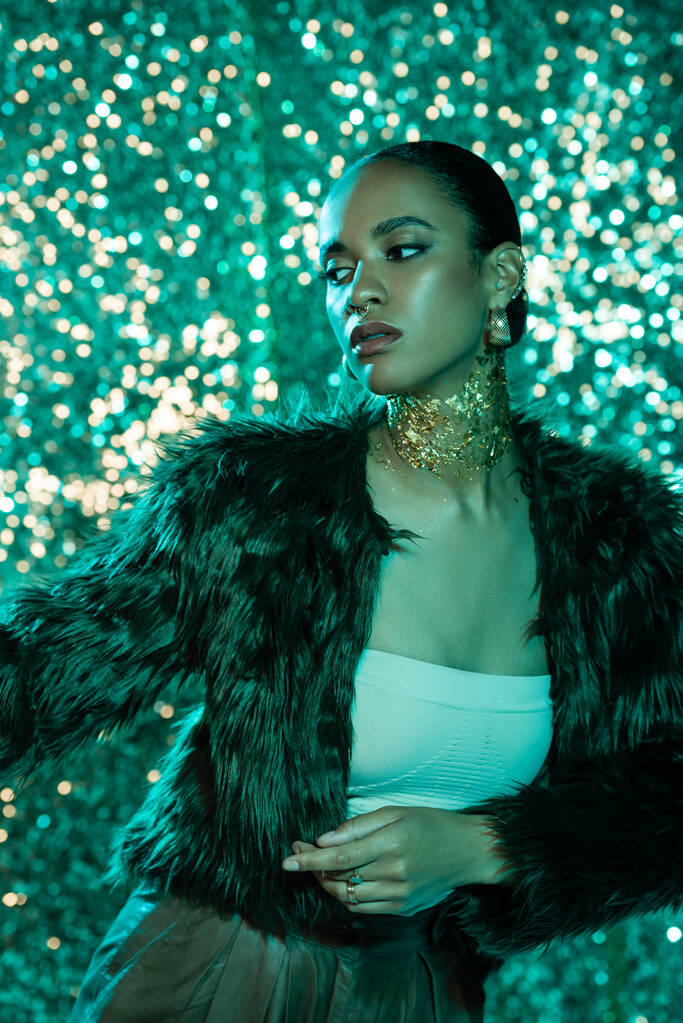 stylowy african amerykański model w faux futro kurtka i złoto na szyi pozowanie na błyszczącym niebieskim tle  - Zdjęcie, obraz