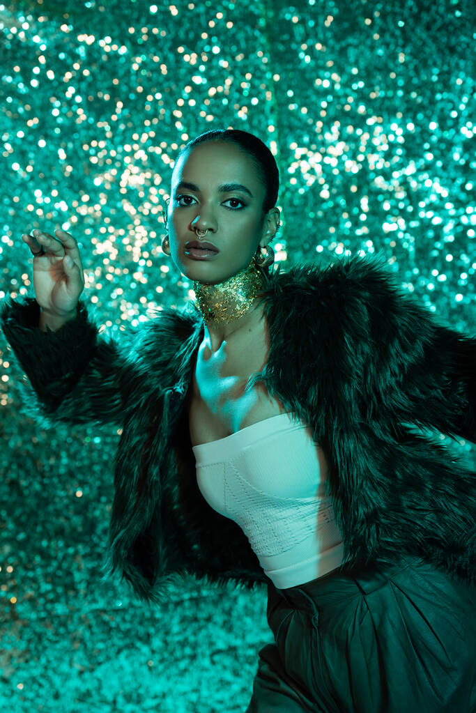 donna afroamericana alla moda in pelliccia sintetica giacca e oro sul collo in posa su sfondo blu lucido  - Foto, immagini