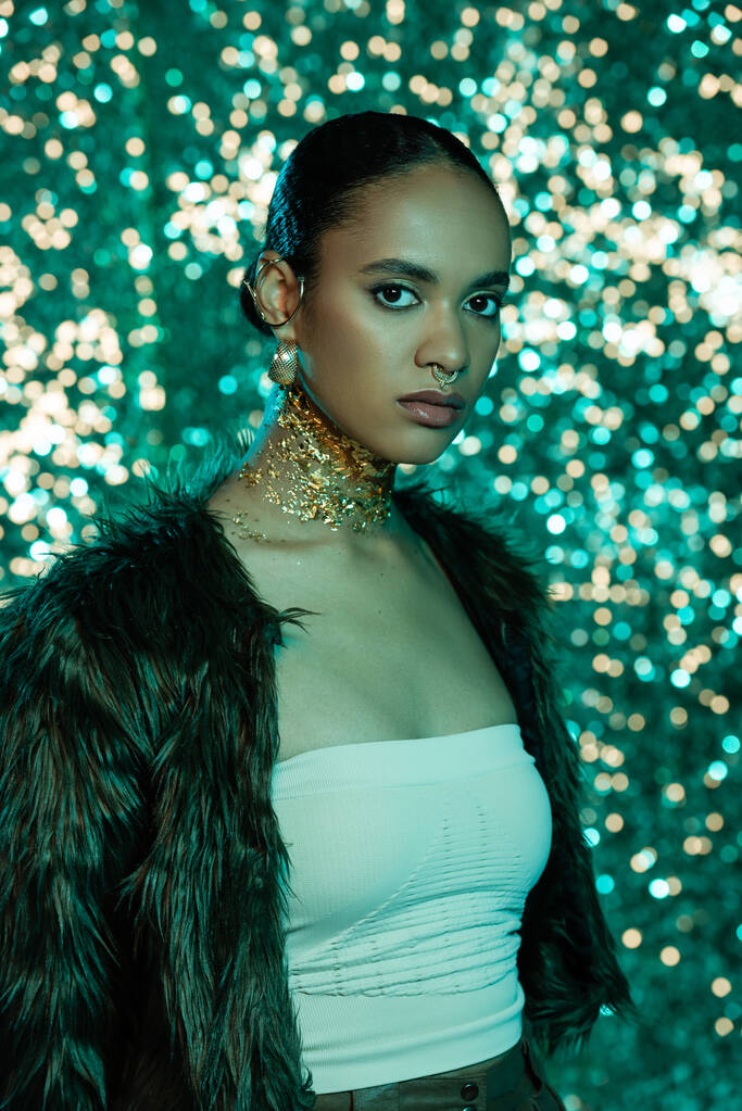 jong Afrikaans amerikaans vrouw in trendy faux fur jas en goud op de hals kijken naar camera op glanzende blauwe achtergrond  - Foto, afbeelding