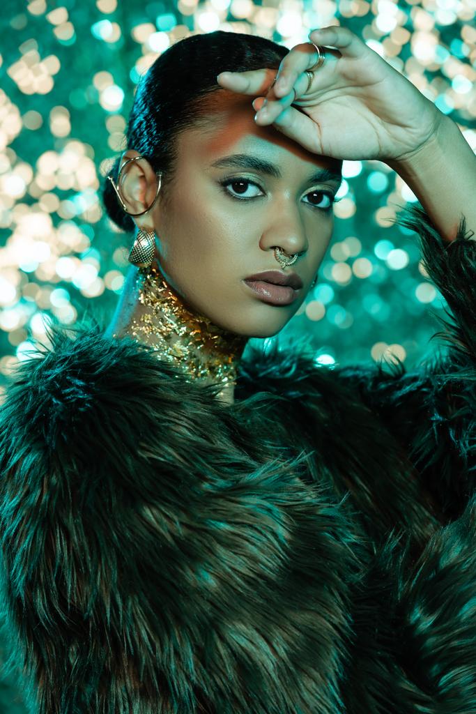 Elegante donna afro-americana in pelliccia sintetica giacca guardando la fotocamera su sfondo scintillante  - Foto, immagini