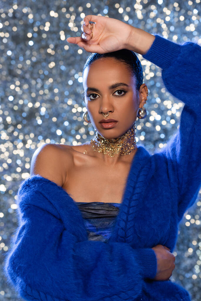 Trendy african american model in blue sweater and golden accessories on sparkling background  - Photo, Image