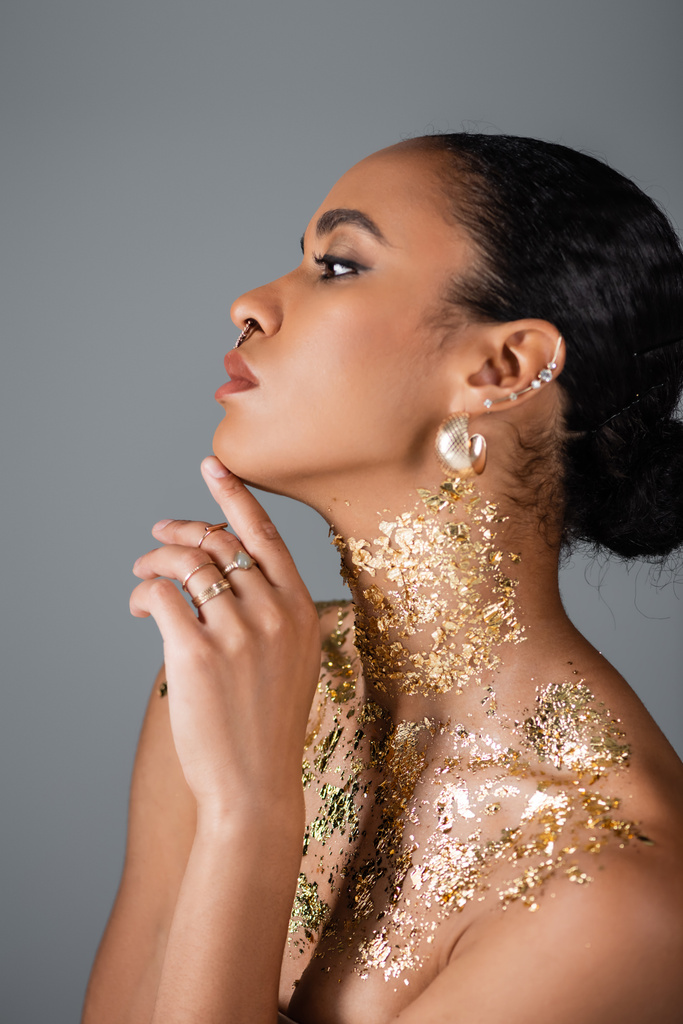Side view of stylish african american model with golden foil on neck touching chin isolated on grey  - Photo, Image