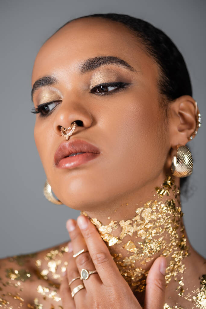 Portrét mladé afroamerické ženy s doplňky a piercing dotýkající se zlaté fólie na krku izolované na šedé  - Fotografie, Obrázek