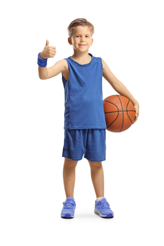 Retrato completo de un niño en un jersey azul sosteniendo una pelota de baloncesto y mostrando los pulgares hacia arriba aislados sobre fondo blanco - Foto, imagen