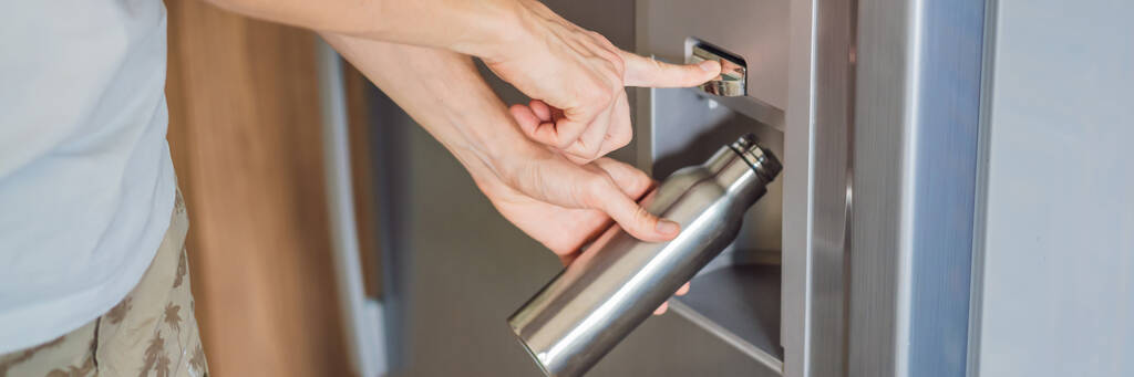 La mano masculina está vertiendo agua fría y cubitos de hielo en una botella de metal del dispensador de la nevera casera. BANNER, FORMATO LARGO - Foto, Imagen