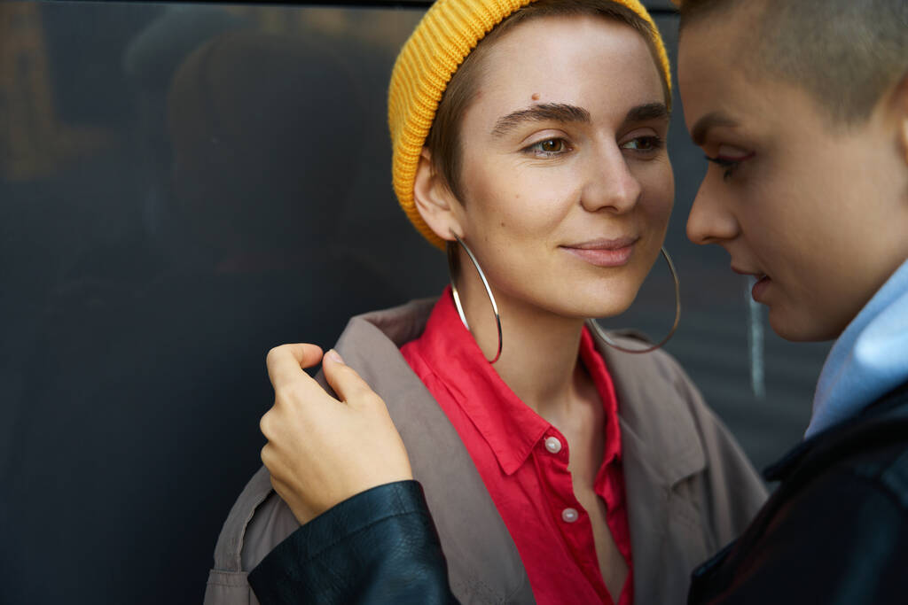 Kadınlar gri bir duvarın önünde birbirlerine yakın dururlar, kadın arkadaşına gülümser, onlar LGBT çiftidir. - Fotoğraf, Görsel
