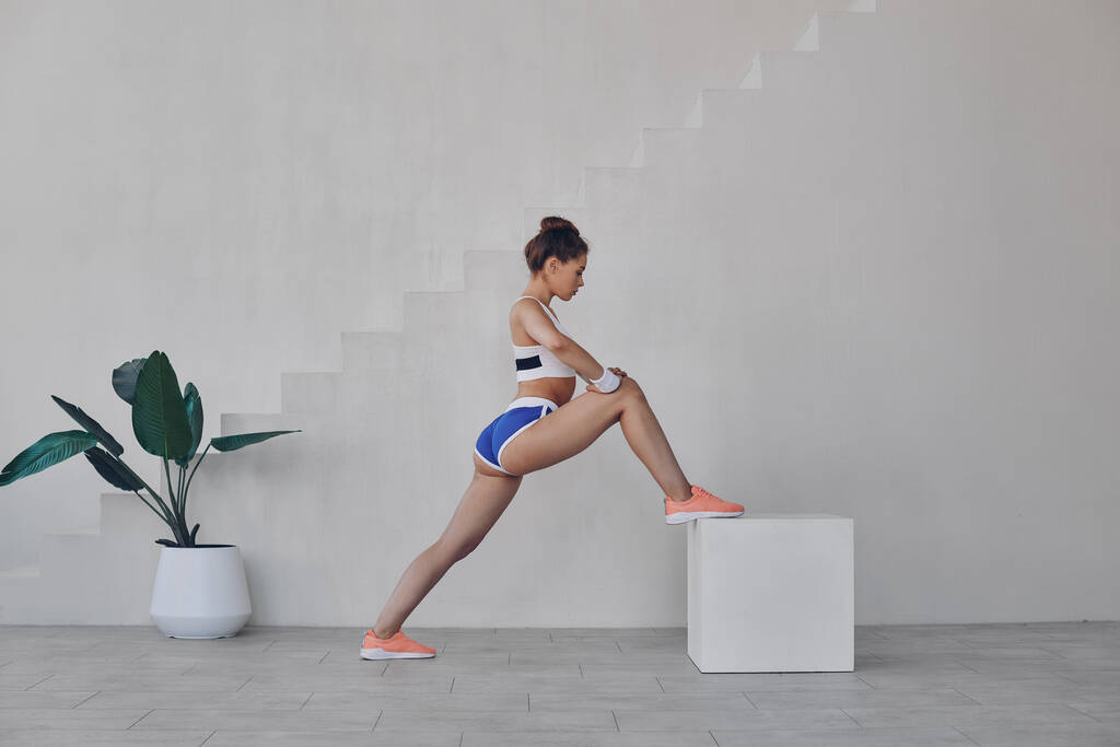 Past jonge vrouw in sport kleding doen stretching oefeningen tijdens het oefenen binnen - Foto, afbeelding