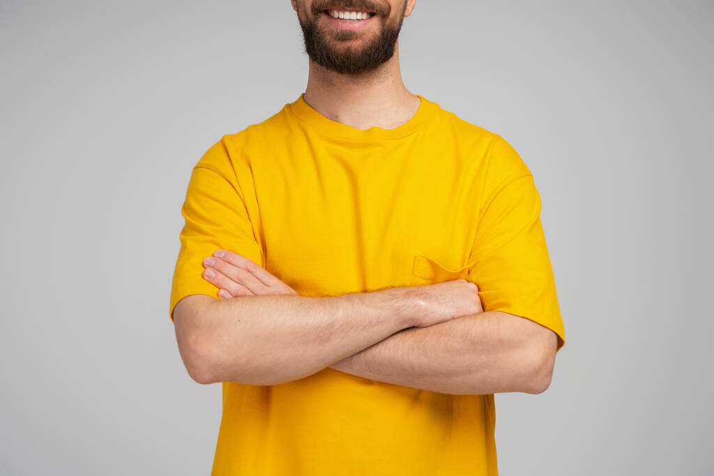 przycięty widok brodatego mężczyzny w żółtej koszulce uśmiecha się stojąc ze skrzyżowanymi ramionami odizolowanymi na szaro - Zdjęcie, obraz