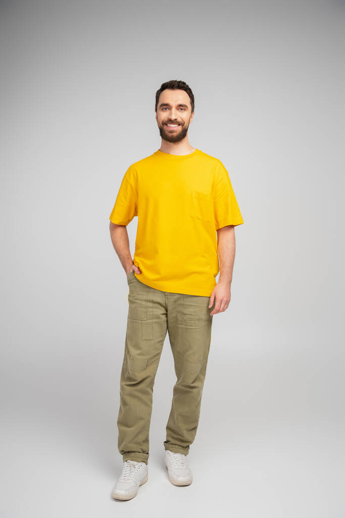 longitud completa de hombre barbudo feliz en pantalones beige y camiseta amarilla de pie con la mano en el bolsillo sobre fondo gris - Foto, Imagen