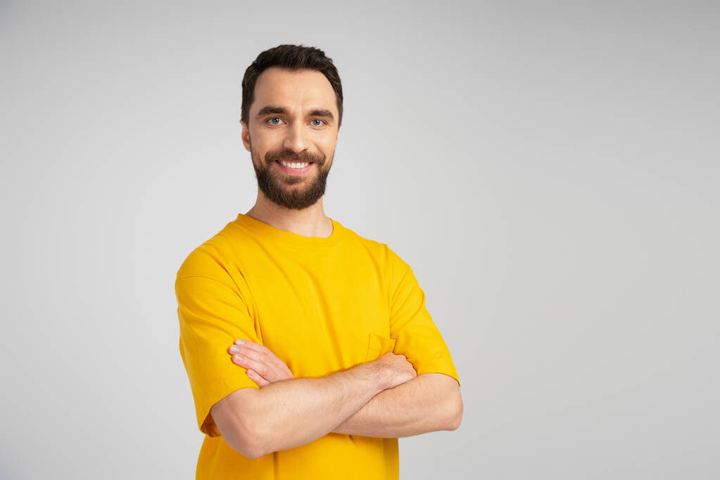 glücklicher bärtiger Mann im gelben T-Shirt lächelt in die Kamera, während er mit verschränkten Armen isoliert auf grau steht - Foto, Bild