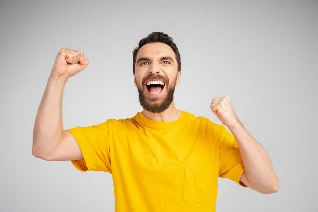 homme excité en t-shirt jaune criant et montrant geste de triomphe isolé sur gris - Photo, image