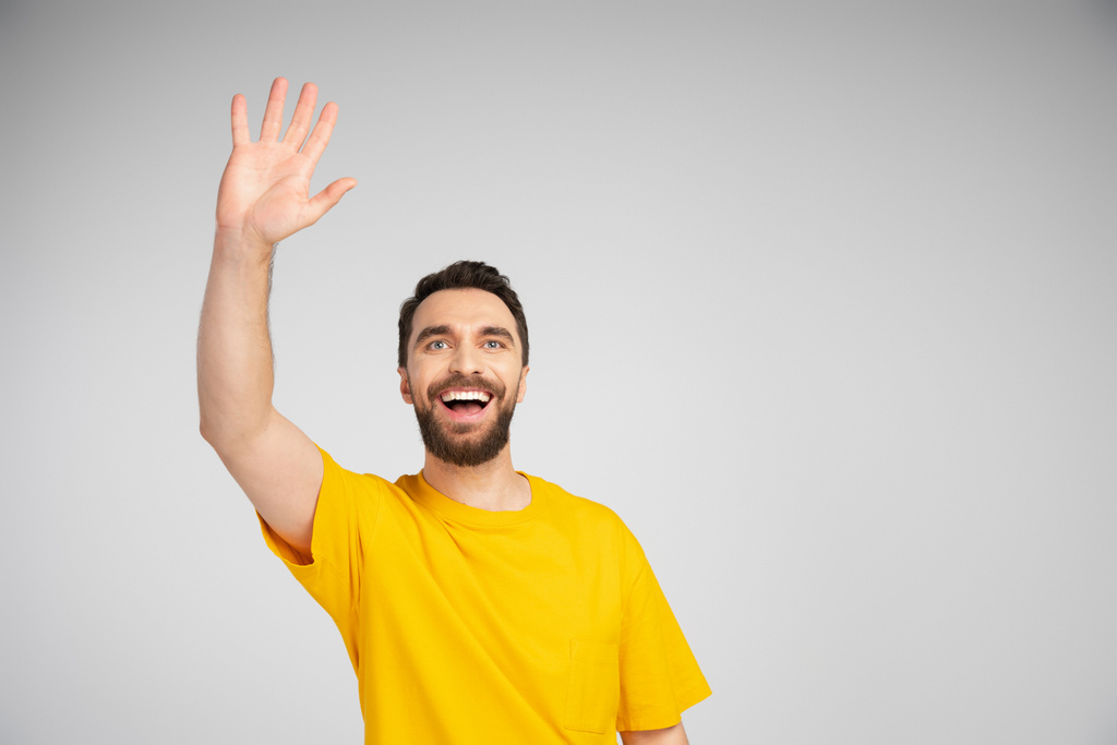Χαρούμενος γενειοφόρος άνδρας με κίτρινο t-shirt κουνώντας το χέρι και κοιτάζοντας μακριά απομονωμένος σε γκρι - Φωτογραφία, εικόνα