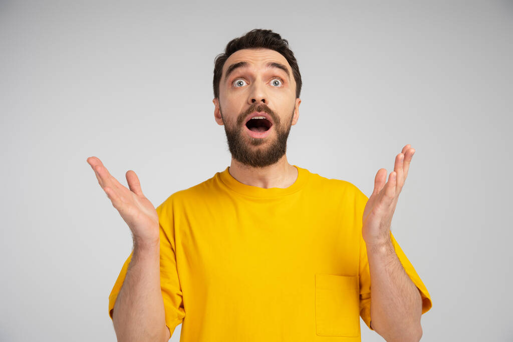 amazed bearded man in yellow t-shirt standing with open mouth and showing wow gesture isolated on grey - Photo, Image