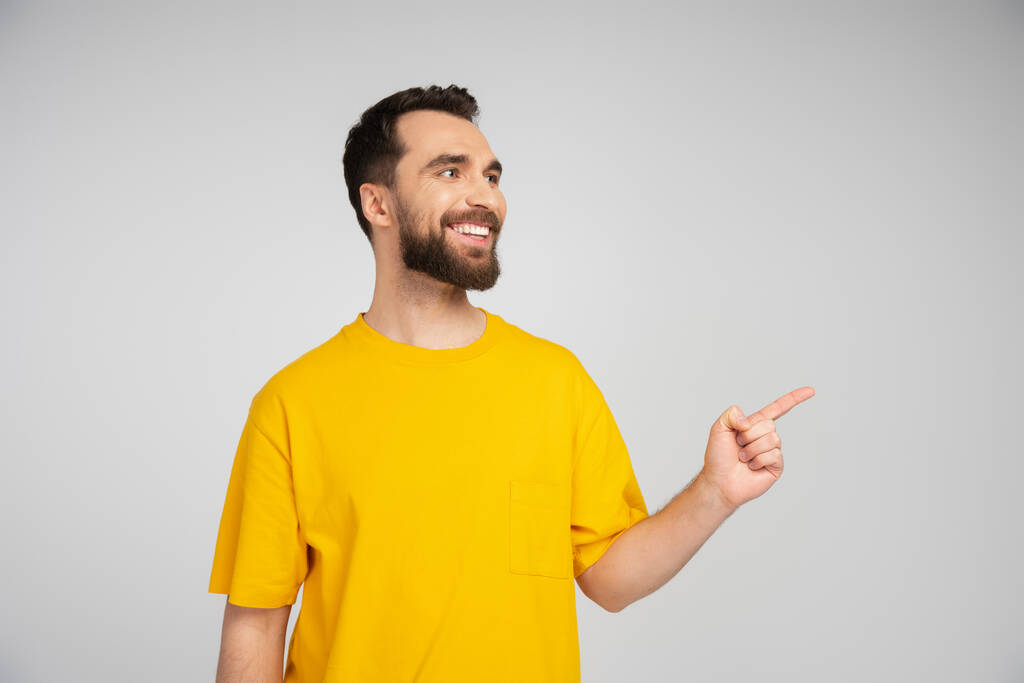 cheerful bearded man looking away and pointing with finger isolated on grey - Photo, Image
