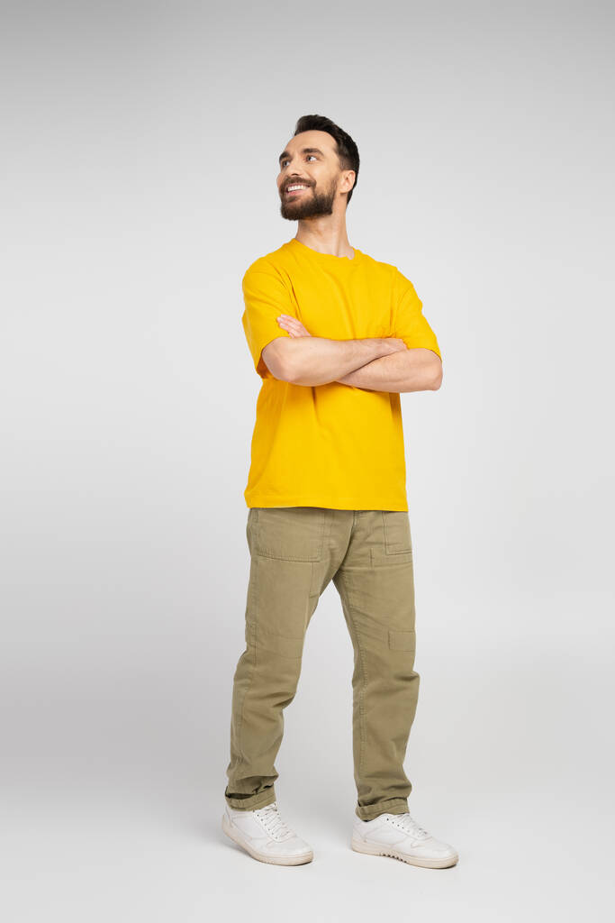 a boldog férfi teljes hossza bézs nadrágban és sárga pólóban, keresztbe tett karokkal és félrenézve a szürkére  - Fotó, kép
