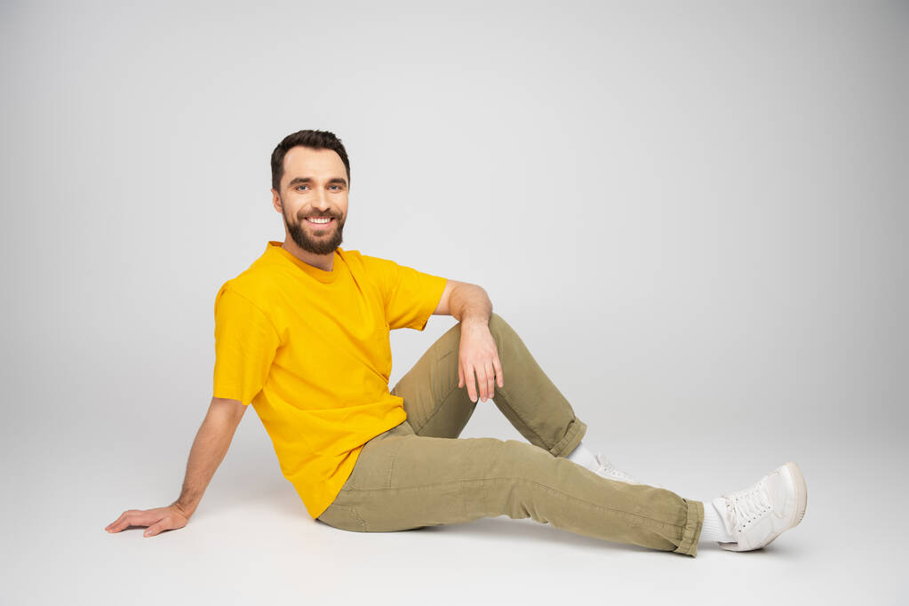 volledige lengte van vrolijke man in beige broek en geel t-shirt zittend op grijze achtergrond - Foto, afbeelding