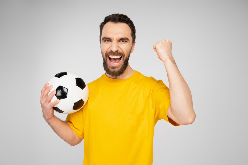 vzrušený vousatý sportovní fanoušek s fotbalovým míčem křičí a ukazuje vítězství gesto izolované na šedé - Fotografie, Obrázek