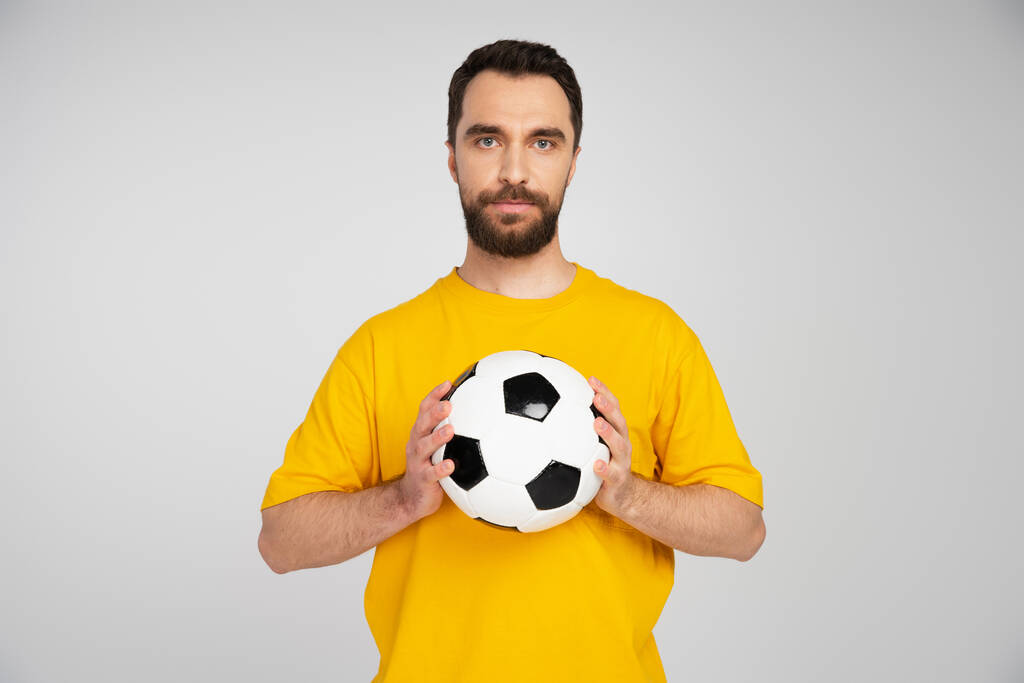 baard sportfan in geel t-shirt houden voetbal terwijl kijken naar camera geïsoleerd op grijs - Foto, afbeelding