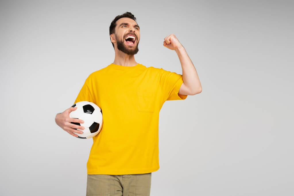 fanático de los deportes llenos de alegría con pelota de fútbol mirando hacia otro lado y mostrando gesto de victoria mientras grita aislado en gris - Foto, Imagen
