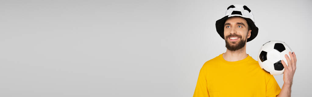 homem feliz em chapéu de fã de futebol de pé com bola de futebol e olhando para longe isolado em cinza, banner - Foto, Imagem
