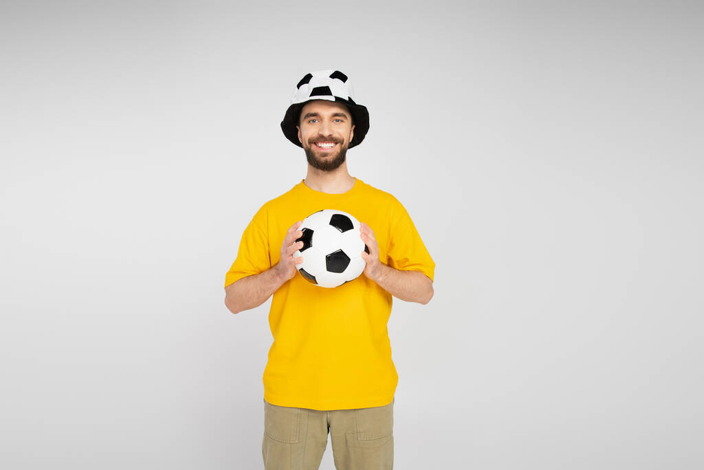 alegre barbudo hombre en fútbol abanico sombrero celebración fútbol pelota y mirando cámara aislado en gris - Foto, Imagen