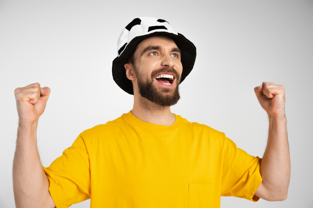 χαρούμενα γενειοφόρος άνδρας στο καπέλο του ποδοσφαιρόφιλου δείχνει κερδίσει χειρονομία και φωνάζοντας απομονώνονται σε γκρι - Φωτογραφία, εικόνα