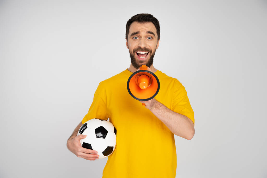 užaslý a šťastný fotbalový fanoušek drží fotbalový míč a křičí v megafonu izolované na šedé - Fotografie, Obrázek